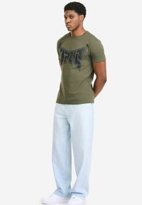 Olivgrünes T-Shirt mit kurzen Ärmeln und einem großen schwarzen "TAPOUT"-Logo, kombiniert mit hellblauen, weit geschnittenen Jeans und weißen Turnschuhen.