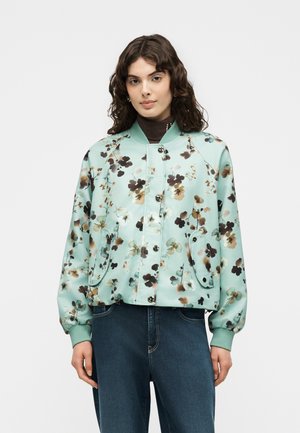 Woman with curly hair wearing a mint green floral bomber jacket over a brown turtleneck and dark blue jeans against a plain background.