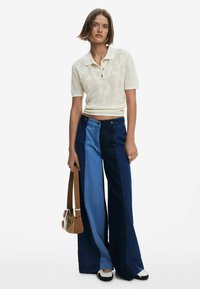 Polo in maglia color crema con motivo floreale, jeans a vita alta e gamba larga in tonalità blu contrastanti, abbinati a una borsa a tracolla marrone e scarpe nere.