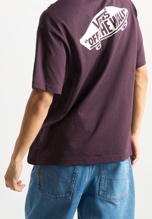 Osoba nosząca ciemnofioletową koszulkę Vans "Off The Wall" i niebieskie dżinsy, pokazana z tyłu na jednolitym tle.