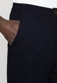 Granatowe spodnie wykonane z gładkiego materiału, z boczną kieszenią i zapięciem na guzik w talii. Prosta nogawka.