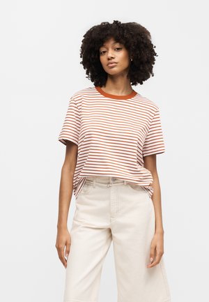 Žena s kučeravými vlasmi, oblečená v bielom a hrdzavom pruhovanom tričku, zastrčenom do krémových širokých nohavíc s vysokým pásom, stojaca pred jednoduchým pozadím.