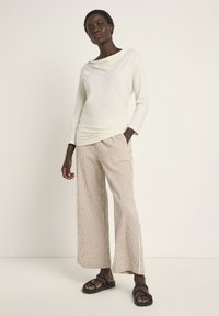 Helle beige Langarm-Bluse mit einem drapierten Ausschnitt, kombiniert mit weiten gestreiften Hosen in Weiß und Terrakotta. Schwarze flache Sandalen.