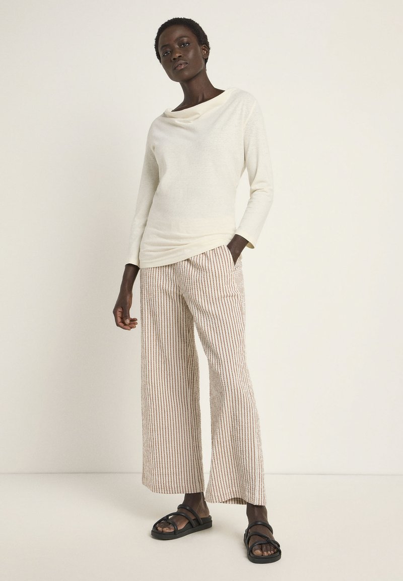 Helle beige Langarm-Bluse mit einem drapierten Ausschnitt, kombiniert mit weiten gestreiften Hosen in Weiß und Terrakotta. Schwarze flache Sandalen.