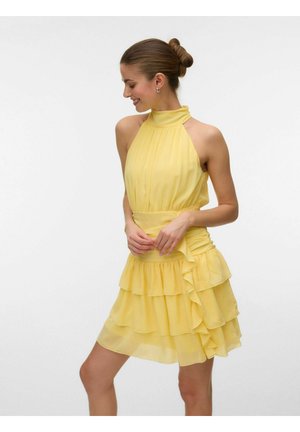 Žena sa usmieva, má na sebe žlté bezrukávové šaty s halterovým výstrihom a vrstvenou volánovou sukňou, vlasy má zaviazané do drdola, stojí pred bielym pozadím.