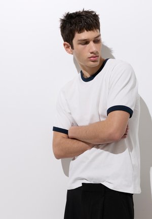 Jeune homme aux cheveux bruns courts portant un t-shirt blanc avec col et bords de manches noirs, les bras croisés, regardant vers le bas à sa gauche.