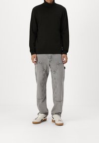 Mężczyzna ubrany w czarny sweter z golfem, szare dżinsy cargo z dużymi kieszeniami i białe trampki z gumową podeszwą, stojący na białym tle.