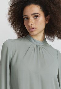 Ljusgrön blus med hög necklinje, veckad detalj vid kragen, långa ärmar och en slät, mjuk textur.