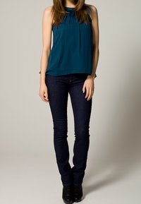 Blouse sans manches bleu sarcelle avec col plissé, associée à un jean skinny foncé et des bottines noires. Tissu lisse et coupe ajustée.