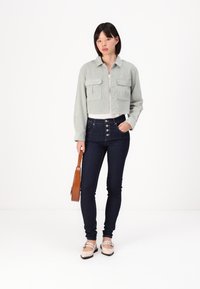 Beskarpet lys grøn jakke med to frontlommer, kombinert med mørkeblå høyt liv jeans og beige leppestifter med dekorative stropper.