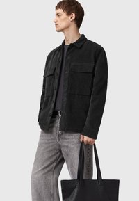 Black corduroy shirt jacket with two chest pockets, worn over a dark top, paired with light gray jeans and a textured black tote bag.