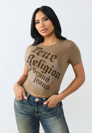 Kobieta z długimi czarnymi włosami, ubrana w dopasowany brązowy t-shirt True Religion i niebieskie jeansy, pozująca z rękami w kieszeniach na jednolitym tle.
