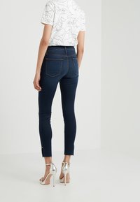 Jeans ajustados oscuros de tiro alto con bolsillos traseros, dobladillo sin terminar con aberturas laterales, combinados con una camiseta gráfica blanca y tacones altos plateados.
