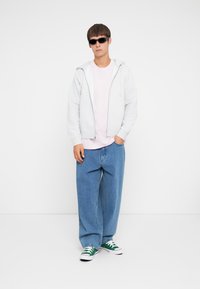Gray zip-up hoodie, light pink t-shirt, loose blue jeans, and green-patterned sneakers. Sunglasses complete the casual outfit.