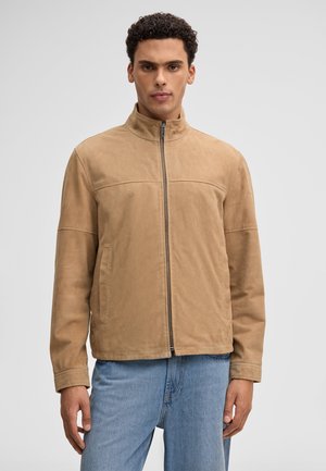 Uomo che indossa una giacca in camoscio color nocciola con zip e colletto alla coreana e jeans azzurri, in piedi davanti a uno sfondo grigio chiaro uniforme.
