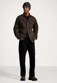 Srajca iz flanela s karo vzorcem v temno zeleni in rjavi barvi, kombinirana s črnimi hlačami in zeleno obutvijo. Model nosi temno pleteno kapo. Prijeten in sproščen kroj.