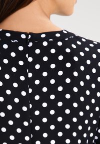 Gros plan sur le cou et l'épaule d'une personne portant un haut noir à pois blancs uniformément espacés avec une petite fermeture éclair noire au dos.