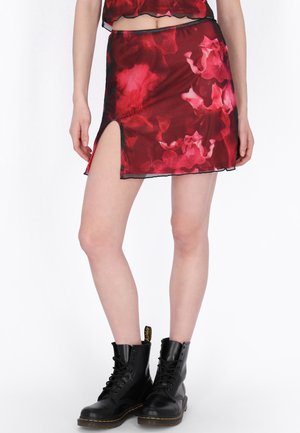 Fustă mini transparentă cu model floral roșu, cu despicătură laterală, purtată cu bocanci negri tip șiret pe un model, pe fundal alb.