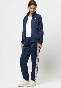 Marineblå treningsdrakt laget av skinnende stoff med hvite og røde Kappa-logo striper langs sidene, med en jakke med glidelås og bukser med elastisk midje.