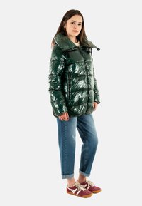 Chaqueta acolchada de un brillante y oscuro verde con un alto cuello, cierre delantero y bolsillos laterales. Combinada con jeans ajustados de un azul claro con puños y zapatillas burdeos.