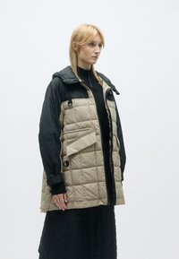 Φουσκωτό μπουφάν δυο τόνων σε μπεζ και μαύρο, με φερμουάρ, κουκούλα και μεγάλη front pocket. Υφαντό ύφασμα με σχέδιο καπιτονέ.