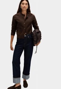 Hnědý pletený cardigan s knoflíkovým zapínáním, dvě přední kapsy; v kombinaci s tmavými džínami s ohrnutými lemy. Hnědé nazouvací boty.