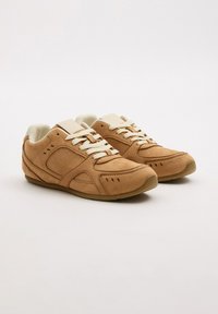 Zapatillas de ante marrón con cordones blancos y una lengüeta acolchada. Presenta paneles laterales curvados y pequeñas perforaciones en la punta. Suela de goma.