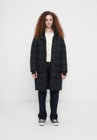 Μαύρο μπουφάν τύπου puffer με κουκούλα, διαθέτει οριζόντιες ραφές, κουμπιά snap και μήκος μέχρι τη μέση. Συνδυάζεται με σκουροκάστανα τζιν και αθλητικά παπούτσια.