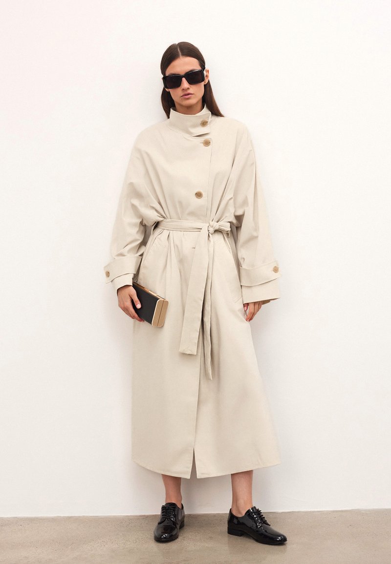 Kvinna i beige bälte trenchcoat, svarta solglasögon, håller en svart och beige kuvertväska, bär svarta blanka oxfordskor, står mot en vit vägg.