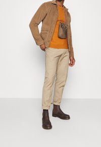 Homme portant une veste en velours côtelé beige, un pull en tricot orange, un pantalon kaki clair, des bottes en cuir brun foncé, et une petite pochette bandoulière marron.