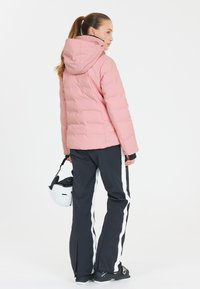Rosa vadderad skidjacka med huva, i kombination med svarta skidbyxor med vita sidostripes. Personen håller en vit skidhjälm.