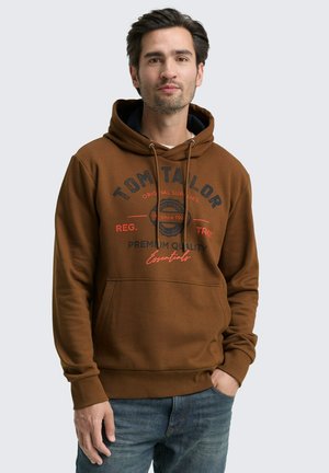 Man in een bruine hoodie met een voorzak en het "Tom Tailor" logo, gecombineerd met blauwe jeans, staande tegen een lichtgrijze achtergrond.