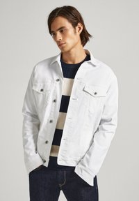 Witte spijkerjacket met twee borstzakken en knoopsluiting, gedragen over een marineblauwe en beige gestreepte trui. Gestructureerde stof, casual ontwerp.