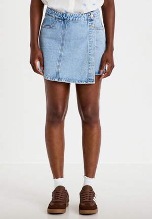 Person standing wearing a light blue denim skirt, white blouse, brown sneakers, and white socks against a plain light background.