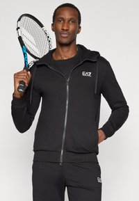 Μαύρο φούτερ με φερμουάρ από απαλό ύφασμα, με ευρύχωρη κουκούλα, μπροστινές τσέπες και λογότυπο EA7. Το μοντέλο κρατά ρακέτα τένις.