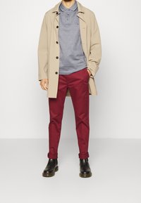 Béžový kabát, modré polo tričko s límcem, bordo kalhoty a černé boty. Tričko má knoflíky a kalhoty mají ohrnuté manžety.