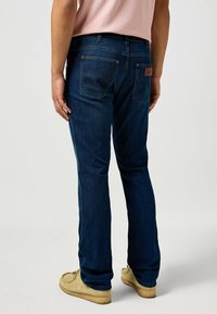 Vaqueros de denim azul oscuro con un diseño de corte recto, que cuentan con bolsillos traseros y un parche de cuero en la cintura. Textura suave, estilo clásico.