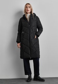 Quiltad svart pufferjacka med huva, med en dragkedja fram, sidofickor och ett diagonalt mönster; bärs över mörka, vida byxor.