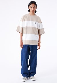 Beige und weiß gestreites, oversized T-Shirt aus Baumwolle, das mit einem kleinen Logodetail versehen ist, kombiniert mit lockeren blauen Jeans und weißen Sneakers.