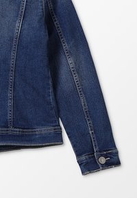 Manica di giacca in denim blu scuro con cuciture visibili e polsino con bottone su sfondo bianco.