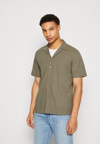 Camisa de botones de manga corta color verde oliva con una tela texturizada, bolsillo en el pecho y cuello clásico, combinada con jeans de color azul claro.