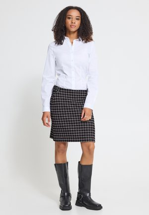 Femme debout portant une chemise blanche boutonnée, une jupe à carreaux noir et blanc, et des bottes noires montantes jusqu'aux genoux sur un fond clair uni.