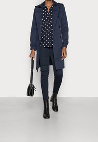 Cappotto trench navy con design doppiopetto, indossato sopra una camicia a pois bianca e nera. Borsa nera e pantaloni scuri aderenti, abbinati a stivali neri.