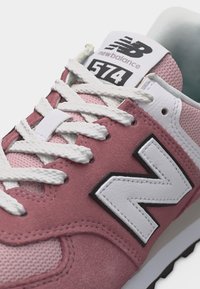 Gros plan sur une basket New Balance 574 rose avec des lacets blancs et un logo sur des matériaux en mesh et en daim.