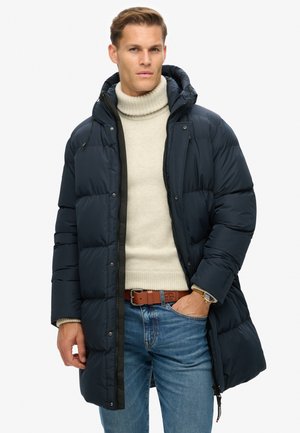 Navy Blousonjacke mit Kapuze, Reißverschluss und gestepptem Design. Getragen über einem cremefarbenen Rollkragenpullover und kombiniert mit blauen Jeans und einem braunen Gürtel.