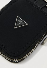 Portefeuille en tissu noir avec une fermeture éclair argentée, présentant une plaque logo triangulaire. La surface texturée et les bords cousus sont visibles.