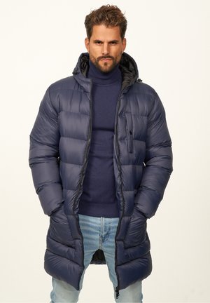 Navy puffer coat featuring a zip front, hood, and elastic cuffs. The quilted design enhances insulation. Worn over a navy sweater and jeans.