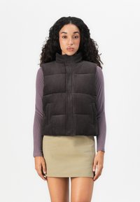 Žena s dlhými kučeravými vlasmi, oblečená v tmavo hnedej nadýchanej veste, pod ktorou má na sebe fialový top s dlhými rukávmi a béžovú sukňu, stojí čelom s rukami pozdĺž tela.