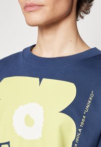 Námořnická mikina se velkým abstraktním žlutým květinovým vzorem, má kulatý výstřih a měkkou texturu. Obsahuje detail s textem "UNIKKO".