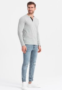 Man med kort brunt hår som bär en ljusgrå långärmad skjorta, slitna ljusblå jeans, vita sneakers och en armbandsur, står upp och ler.